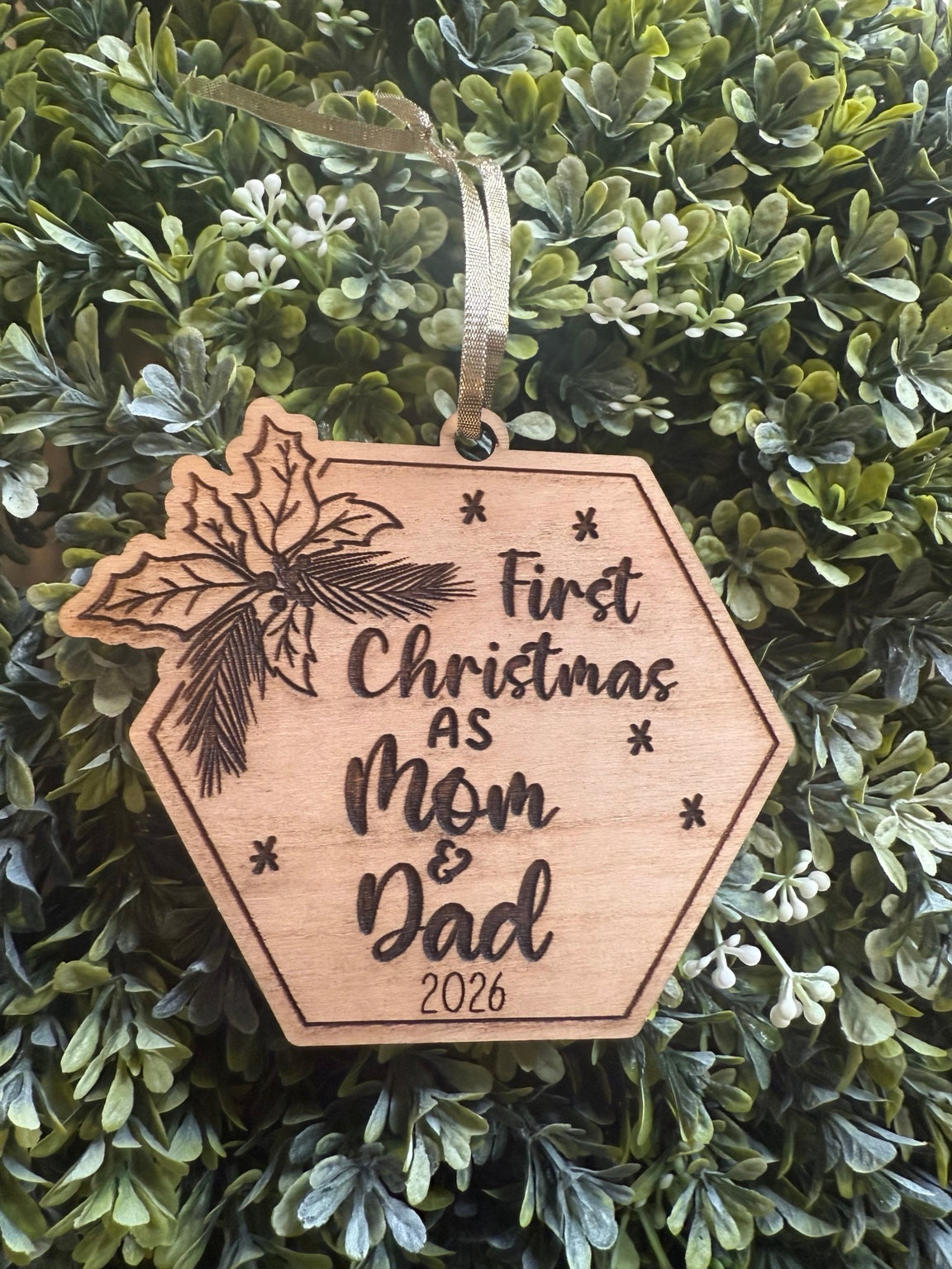 Holly Jolly wooden ornament engraved First Christmas as Mom & Dad 2026, with holly leaves and gold ribbon on greenery